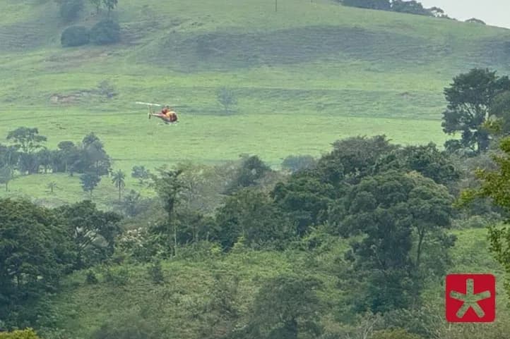 Helicóperto sobrevoando aéra de mata verde, com morros ao fundo