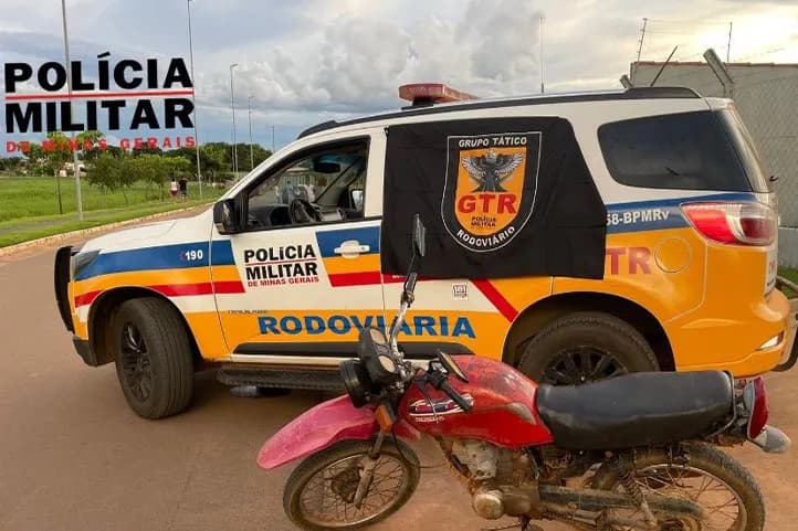 imagem colorida, contendo uma viatura da PM ao lado da motocicleta vermelha, estacionadas ao lado a rodovia