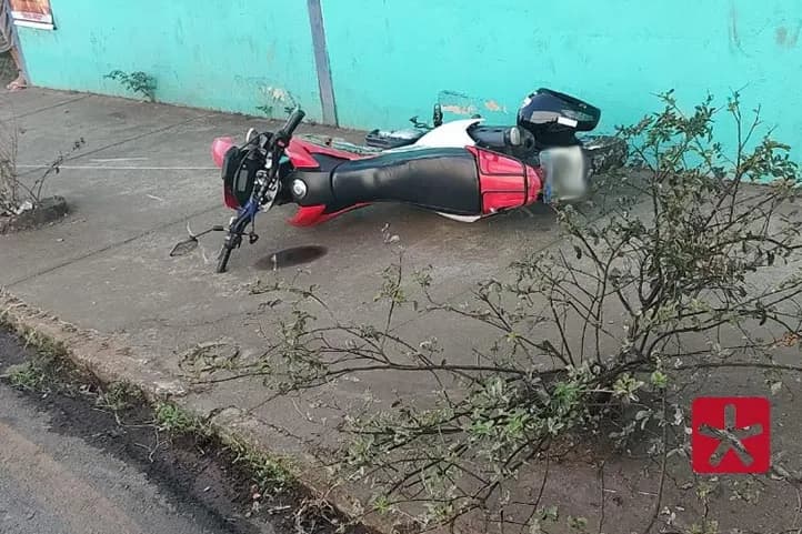 imagem colorida mostrando motocicleta nas cores vermelha, preta e branca caida sobre a calçada. Ao lado uma planta e uma parede azul. 
